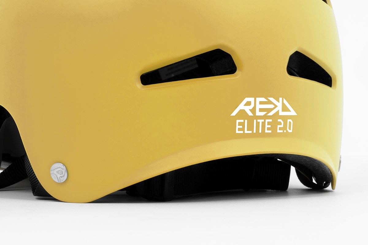 Close-up of the REKD Elite 2.0 Skate / Scooter Helmet in grey and yellow, featuring black vents, "REKD ELITE 2.0" in white on the side, and a round silver emblem near the edge highlighting its certified safety status.