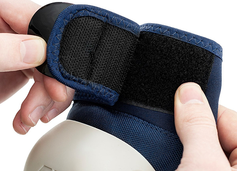 Close-up of hands fastening a black Velcro strap on the REKD Shield Triple Skate Protection Pad Set in blue, with a gray rounded section visible at the bottom.