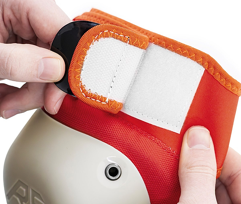 Close-up of hands fastening a white and orange Velcro strap on a REKD Shield Triple Skate Protection Pad, part of the Fade Orange / Green set designed for action sport protection on knees, elbows, or wrists.