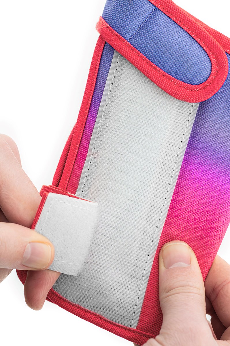 A person opens a REKD Shield Triple Skate Protection Pad from the Fade Red / Violet set, using their hands to undo the white Velcro strap, similar to fastening wrist guards before action sports. The pad features a striking gradient of red, purple, and pink fabric with blue trim at the top.