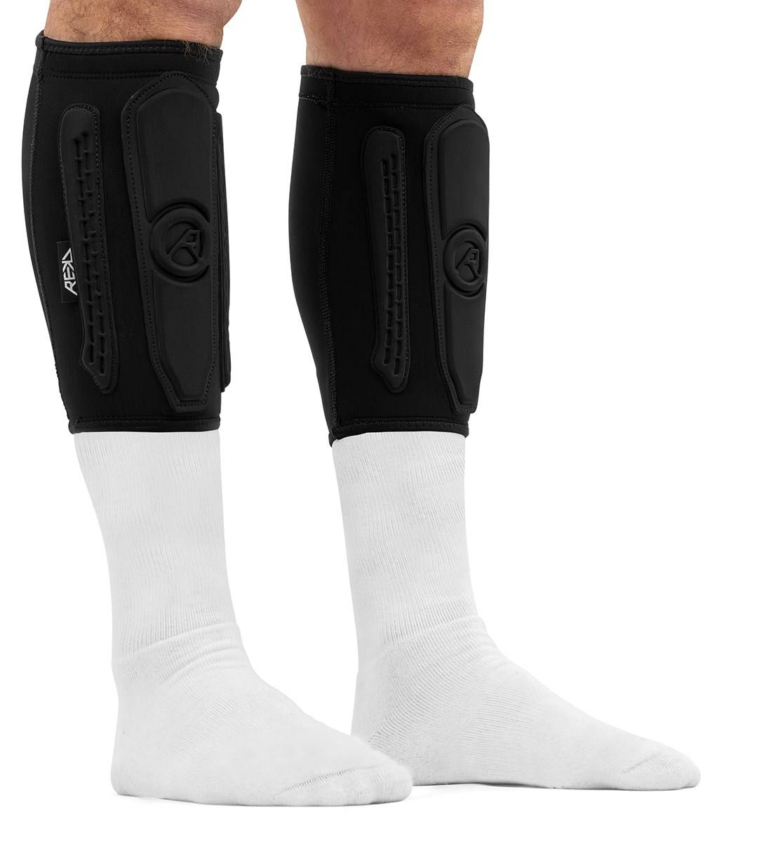 A pair of black REKD Energy Covert Short Shin Skate Protection Guards worn over white socks on a person’s lower legs for slim impact protection.