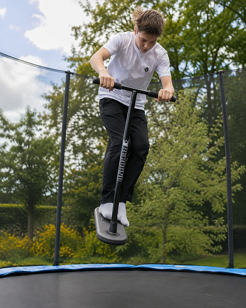 A boy in a white shirt and black pants jumps on a Legacy Rebound Indoor Trampoline Scooter – Electro Black by Legacy, using a Unijump stick, surrounded by green trees and a bright, partly cloudy sky as he showcases his skills.