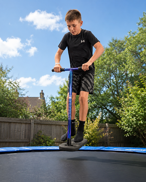 A boy in a black t-shirt and shorts jumps on a Legacy Rebound Indoor Trampoline Scooter - Blueray by Legacy on a trampoline in a backyard, surrounded by greenery, a fence, and under a partly cloudy sky.