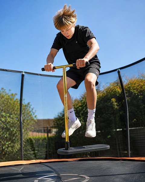 A boy in black athletic clothes jumps on the Legacy Rebound Indoor Trampoline Scooter - Electro Gold by Legacy, enjoying a pogo stick-like ride against a backdrop of blue sky and greenery—a perfect snapshot of the product’s thrilling experience.