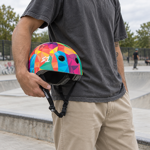 A person holding a multicolored geometric S1 Lifer Skate / Scooter Helmet - Kaleidoscope at a skatepark.