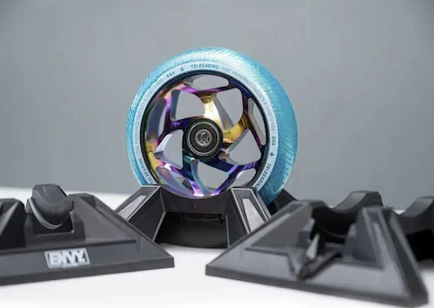 A colorful metallic scooter wheel with a blue tire and 30mm wide wheels stands upright in a Blunt Envy Scooter Stand V2 - Black on a white surface with a gray background.