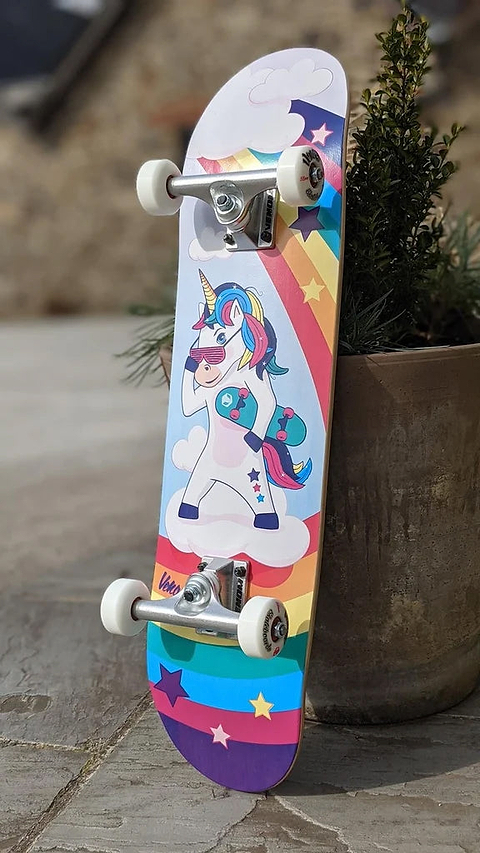 Venom Junior Unicorn Kids Complete Skateboard - 7.25″ x 29″ featuring a cartoon unicorn wearing sunglasses and carrying a smaller skateboard, set against a background of clouds, stars, and a rainbow.