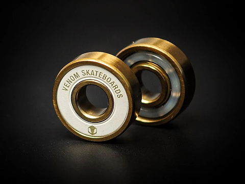 Two gold-colored Venom Precision Gold Skateboard Bearings with white central rings, each labeled with “Venom Skateboards.”