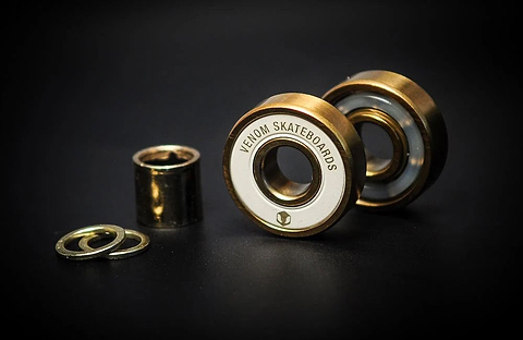 Two gold-toned Venom Precision Gold Skateboard Bearings, a cylindrical spacer, and two washers on a black surface.