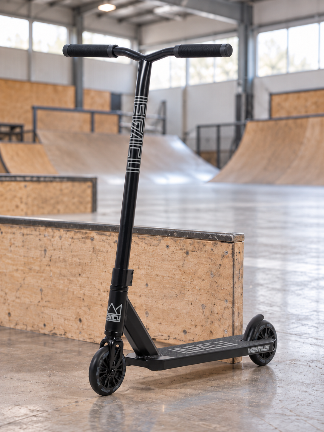 The Legacy Ventus Complete Stunt Scooter - Black from Legacy stands on the concrete floor of an indoor skatepark, with wooden ramps and railings visible in the background—making it the perfect pro scooter for tackling tricks and stunts.