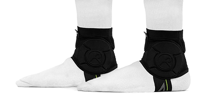 Two feet wearing white socks and black REKD Energy Covert Ankle Impact Skate Protection Guards are shown from the side. The REKD guards feature energy-absorbing circular padding and a logo, set against a plain white background.
