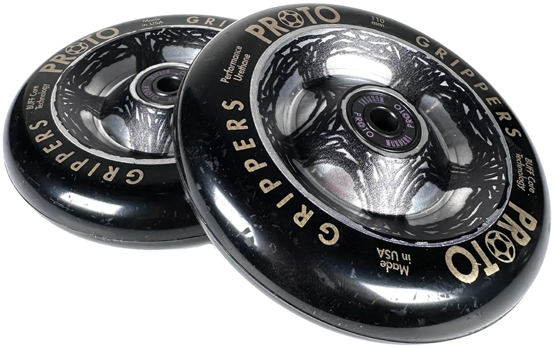 Two black Proto Catalyst Gripper 110mm stunt scooter wheels by Proto, featuring silver and gray graphics plus "PROTO GRIPPERS" and "Performance Urethane" text on the outer edge, are stacked with a slight overlap.