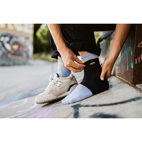 Wearing REKD’s Energy Slim Footies Ankle Protection Pad in black over light blue socks, a person sits outdoors with one bare foot and one in a white sneaker, blending urban style with extra ankle protection amidst graffiti surroundings.