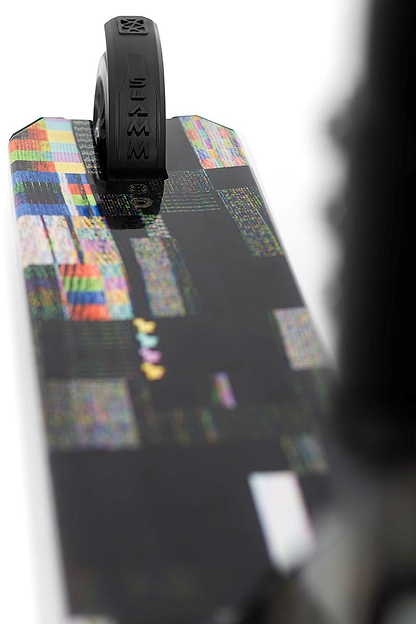 Close-up of a skateboard deck with black grip tape at the tail, displaying pixelated colorful patterns like a nebula wrap. A black wheel is at the top, suggesting agility like the sleek design of the Slamm Mischief Complete Stunt Scooter - Glitch.