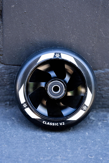 The Union Classic V2 110mm Stunt Scooter Wheel - Black by Union, featuring white markings, stands upright against a dark, textured wall and ground.