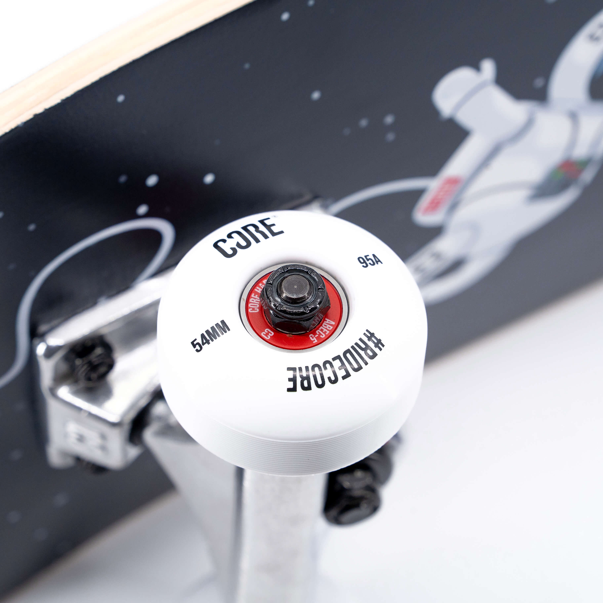 Close-up of a white CORE C1 skateboard wheel with text details and a red bearing, mounted on a silver truck against a deck featuring a spaceman illustration on a CORE C1 Spaceman Complete Skateboard - 7.75″ x 31″.