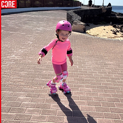 Wearing the CORE Junior Triple Skate Protection Pad Set in pink by CORE, a young child smiles as they skate along a sunlit brick pathway near a rocky beach, protected by their matching helmet, knee pads, and roller skates.