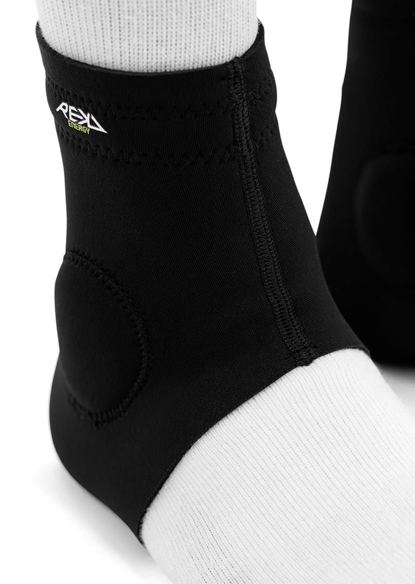 Close-up of feet in white socks and REKD Energy Slim Footies Ankle Protection Pads (Black) with a small "REKD" logo. These pads provide compression and impact protection for the ankle area, ideal for active use.