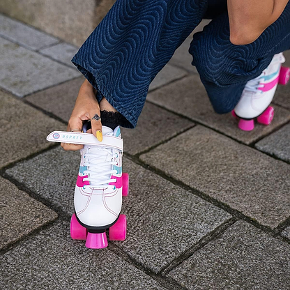 Wearing blue wavy-patterned pants, a person kneels on a stone pavement and adjusts the strap of Osprey Disco Quad Roller Skates - White with bright pink wheels, ready for fun in these stylish Osprey recreational skates.