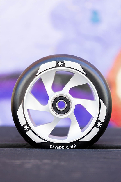 A close-up of the Union Classic V2 110mm Stunt Scooter Wheel in Black/Silver by Union, featuring "CLASSIC V2" text, upright on a dark surface with a blurred, colorful background.
