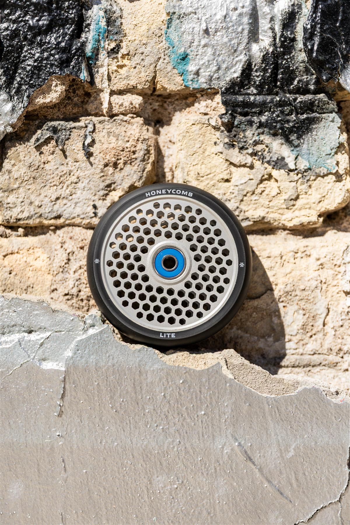 A Union Honeycomb Lite 110mm Stunt Scooter Wheel in black and silver, featuring a round metal honeycomb pattern and blue center, rests against a weathered brick wall.