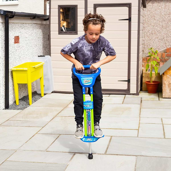 A young child with curly hair jumps on an Ozbozz Go Light Up Kids 89cm Pogo Stick - Blue in a paved backyard near a yellow bin and beige garden shed.