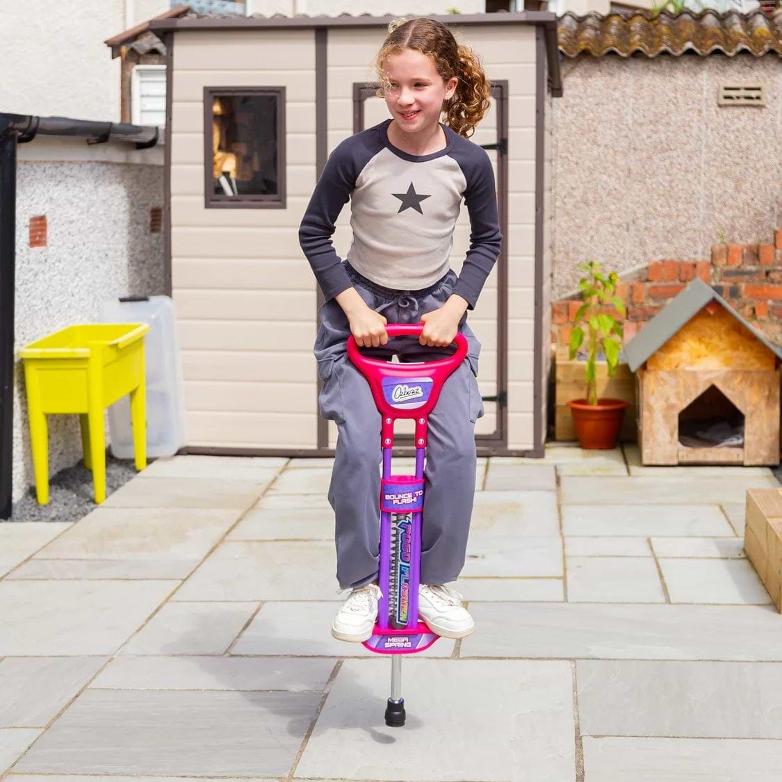 A young girl with curly hair, wearing a gray star sweater and gray pants, bounces on an Ozbozz Go Light Up Kids Pogo Stick - Pink by Ozbozz in a sunny backyard with paved stones and a small shed in the background.