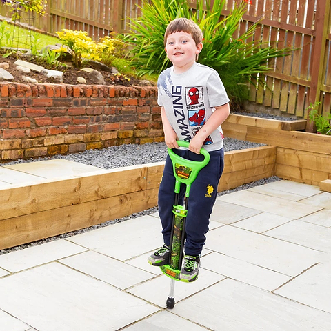 A young boy in a Spider-Man T-shirt and navy pants smiles while jumping on the Ozbozz Dinosaur Kids Pogo Stick - Green by Ozbozz, on a backyard patio with stone tiles and wooden fences.