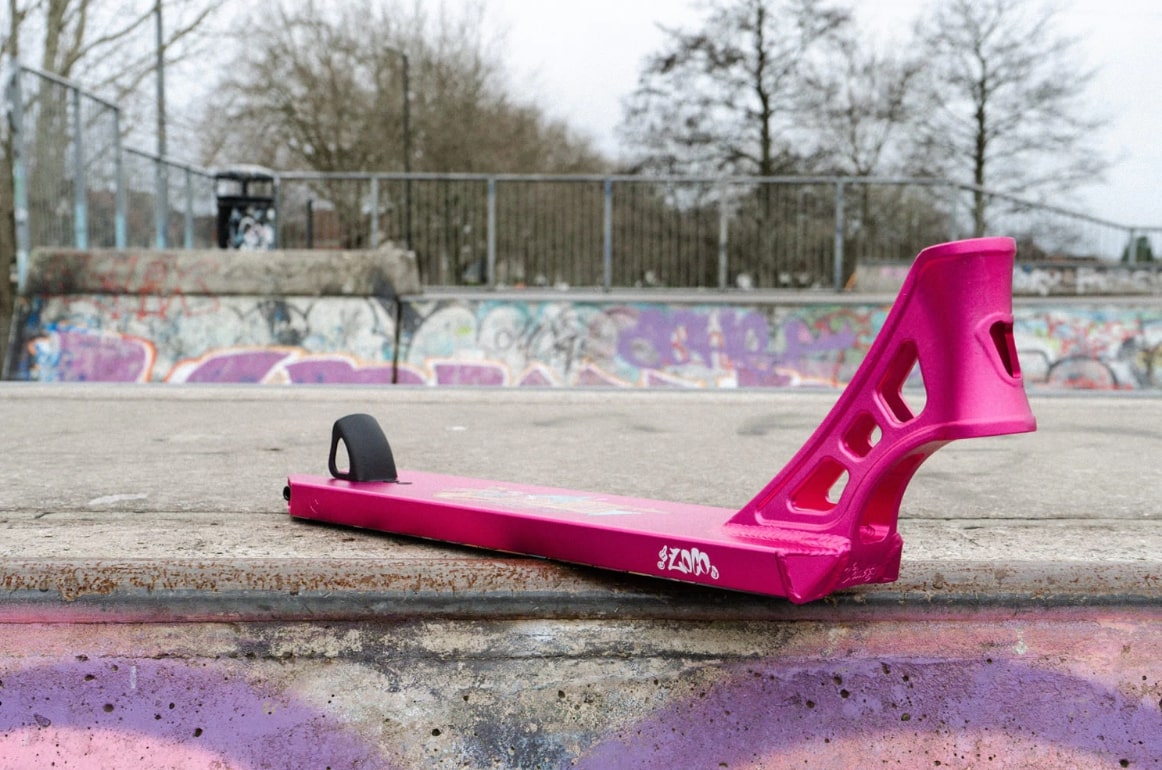 A Chubby Loco Doughnut King Pink Stunt Scooter Deck, measuring 4.5" x 19.5", is positioned on a concrete surface at the skate park, surrounded by graffiti-covered walls and leafless trees.