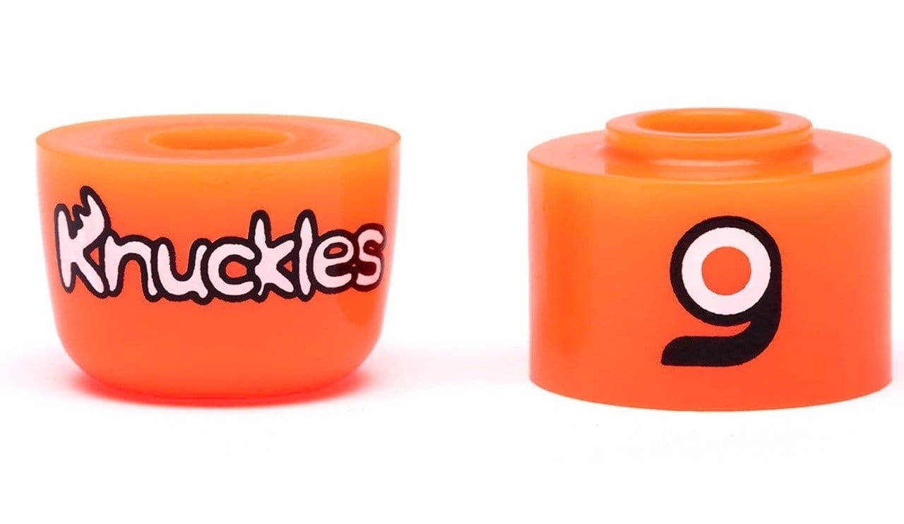 The image showcases two skateboard bushings in orange. The left bushing prominently displays the product name, "Orangatang Knuckles Orange Longboard Bushings - Soft 87A," emphasizing its high-rebound formula. The right bushing is adorned with a stylized black and white "9" logo, adding a sleek touch to these insert barrel bushings from Orangatang.