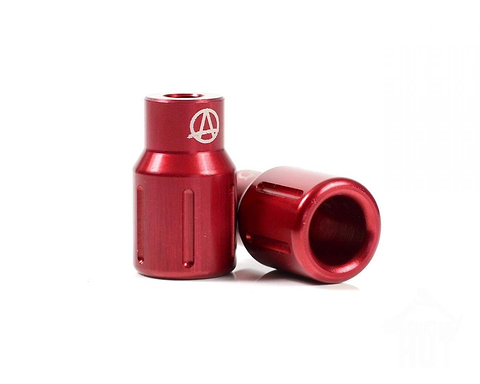 Two red anodized valve caps are displayed, one positioned upright and the other resting against it, reminiscent of the Apex Barnaynay Aluminium Stunt Scooter Pegs. Each features a smooth, metallic finish with a white engraved logo on the top of the upright cap.