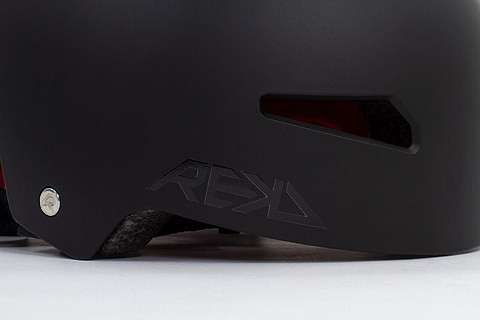 Close-up of the black REKD Elite 2.0 Skate/Scooter Helmet featuring the "REKD" logo and a ventilation slot. This certified action sports helmet highlights a metallic screw detail on the side, with its interior lining visible through the ventilation opening.