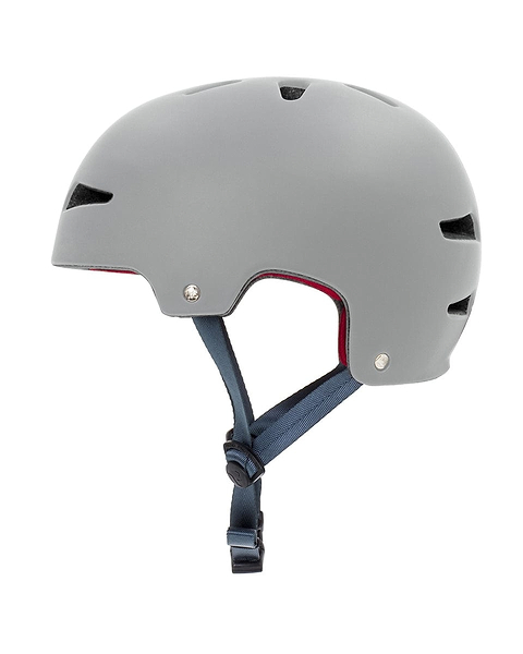A side view of the REKD Ultralite In-Mold Skate/Scooter Helmet in grey highlights its matte finish. This helmet, designed for action sports, includes ventilation slots on the top and sides. It also features a blue adjustable chin strap with a black buckle. The advanced In-Mold technology is visible through the red interior lining, offering improved safety.