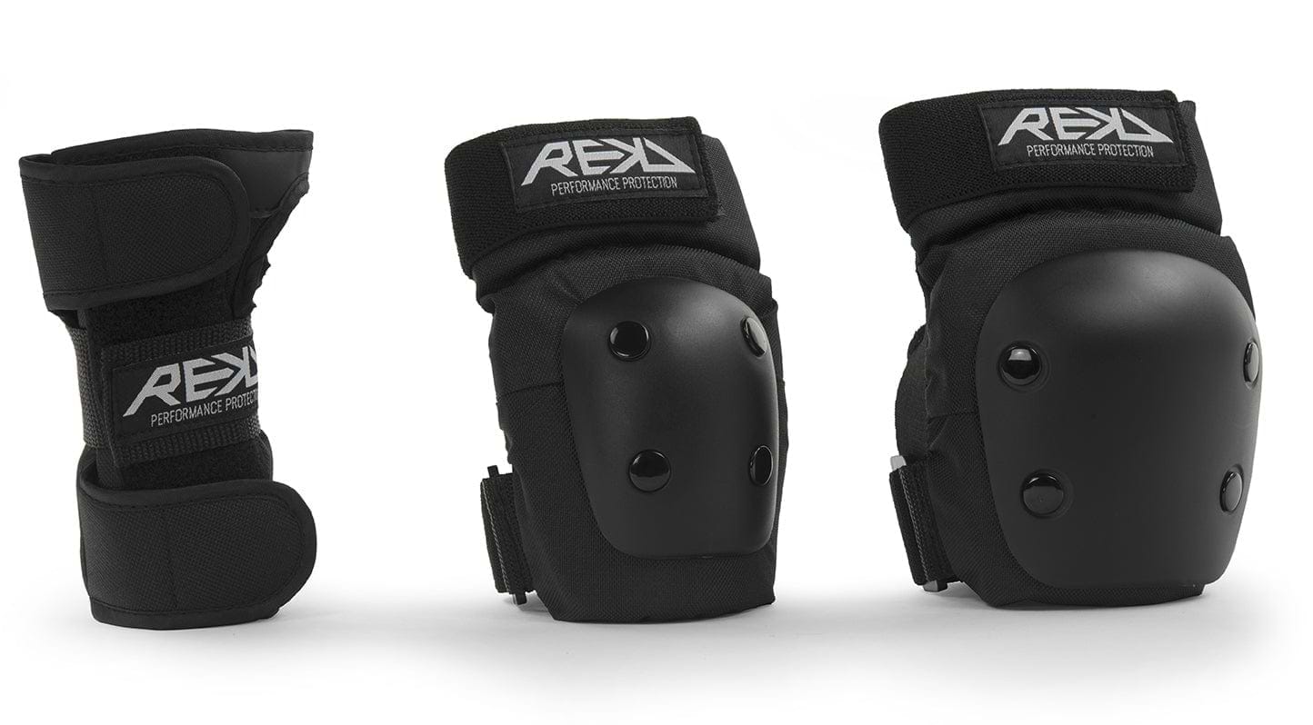 Three black protective gear items from REKD are showcased: an ergonomic wrist guard on the left and two knee or elbow pads on the right. The REKD Junior Heavy Duty Triple Skate Protection Pad Set includes hard plastic shells with the "REKD" logo on the straps, providing protection for extreme sports enthusiasts.