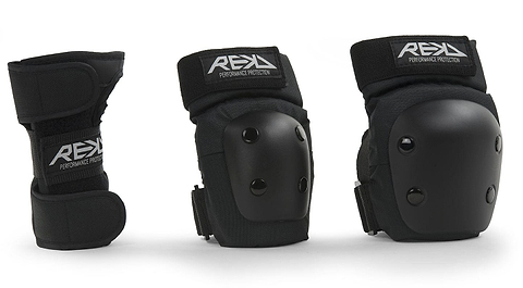 Three black protective gear items from REKD are showcased: an ergonomic wrist guard on the left and two knee or elbow pads on the right. The REKD Junior Heavy Duty Triple Skate Protection Pad Set includes hard plastic shells with the "REKD" logo on the straps, providing protection for extreme sports enthusiasts.
