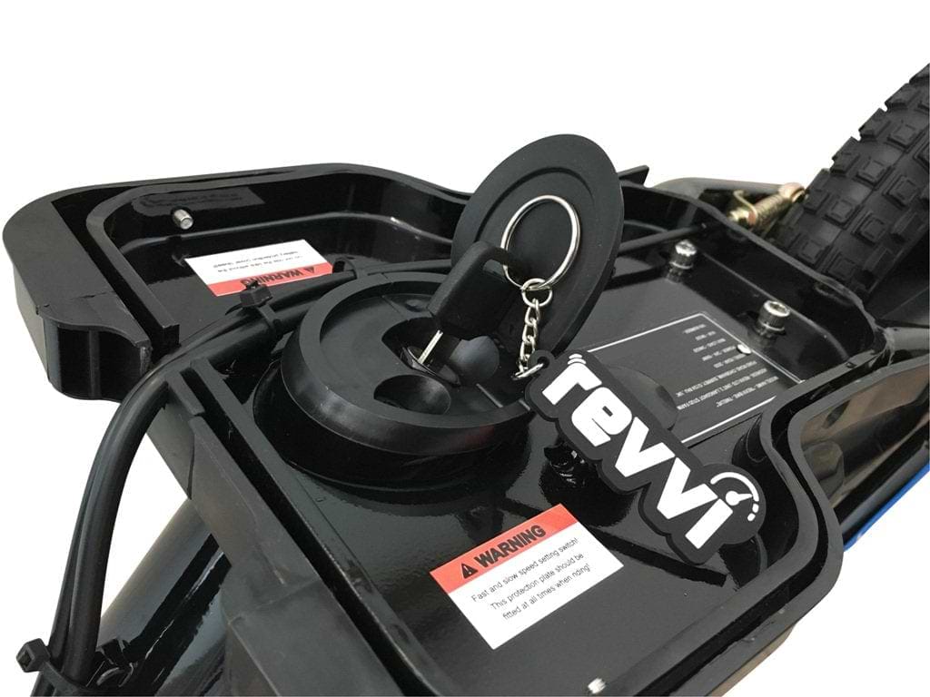 Close-up of the Revvi 16" Plus Kids Electric Balance Bike in orange, highlighting its charging port with a key attached to a "revvi" keychain label. A warning label is seen nearby, with the bike's tire and rear section visible in the background.