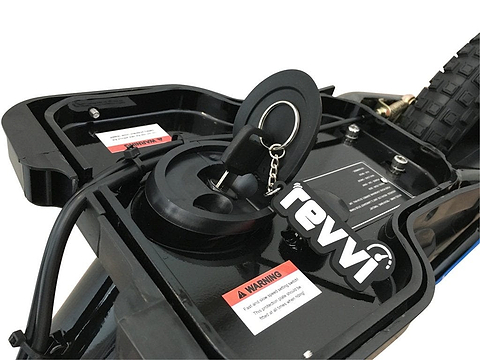 Close-up of the Revvi 16" Plus Kids Electric Balance Bike in orange, highlighting its charging port with a key attached to a "revvi" keychain label. A warning label is seen nearby, with the bike's tire and rear section visible in the background.