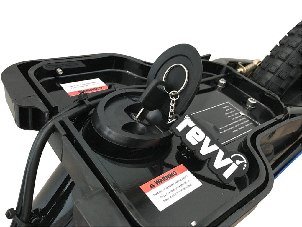 The image shows a close-up of a black battery compartment on an electric bike. A key is inserted into the lock, attached to a keychain labeled "Revvi 16\" Plus." In the background, a tire suggests that this could be the Revvi 16" Plus Kids Electric Balance Bike in pink, with warning labels visible on the battery case.