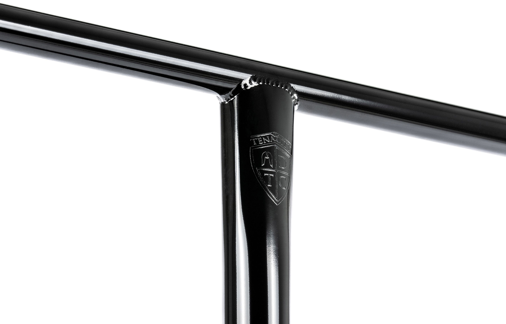 Close-up of a glossy black metal T-joint featuring a small engraved shield logo reading “TENN” on the vertical bar, showcasing the exceptional craftsmanship of the Ethic DTC Tenacity V2 Steel Oversized HIC Stunt Scooter T-Bars in Black Mirror (770mm x 600mm). Manufactured by Ethic, these oversized chromoly steel bars deliver outstanding strength and durability for demanding riders.