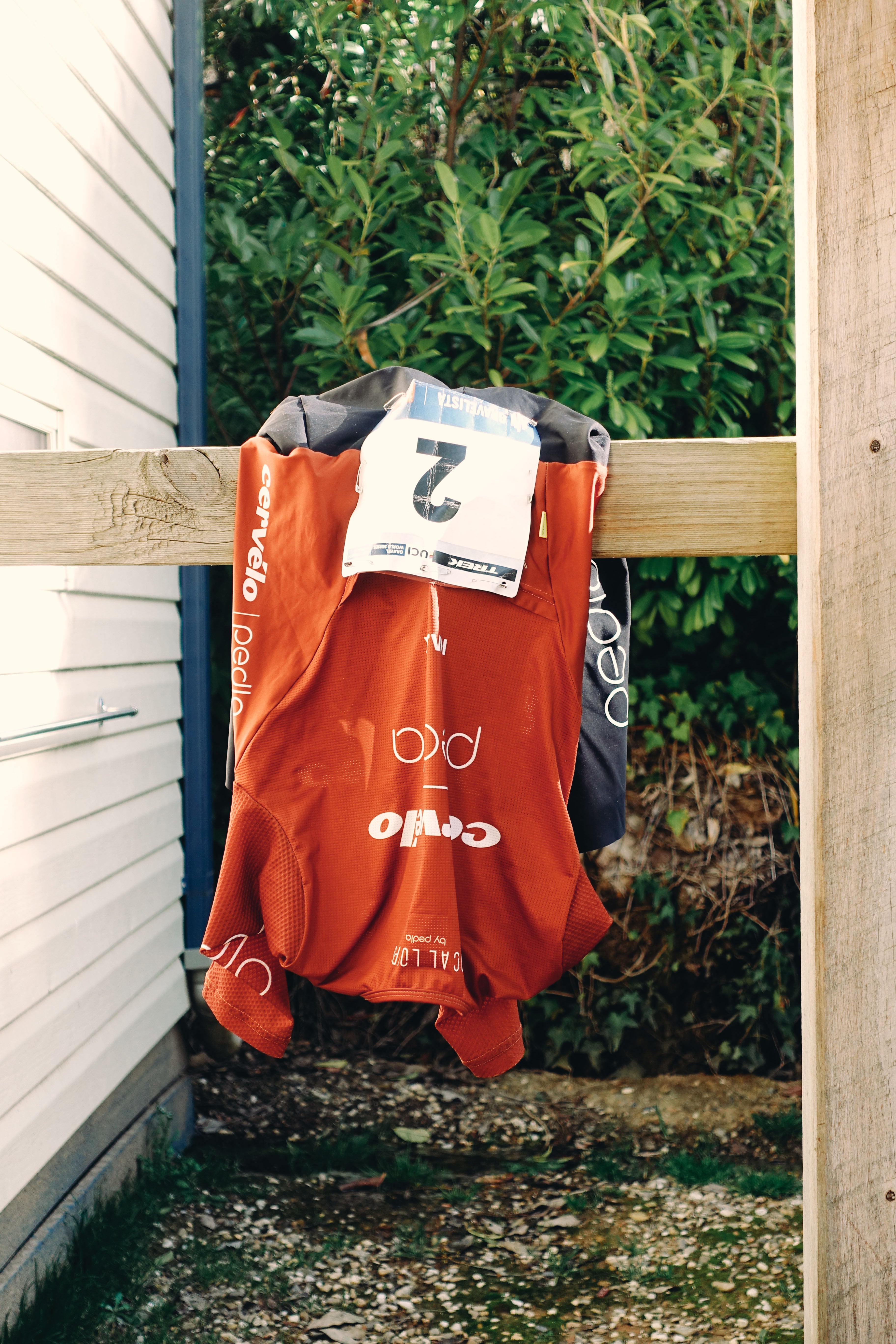 Image of the Cervelo Pedla race jersey hanging on a railing, with a race number pinned on. The jersey is bronze with white logos.