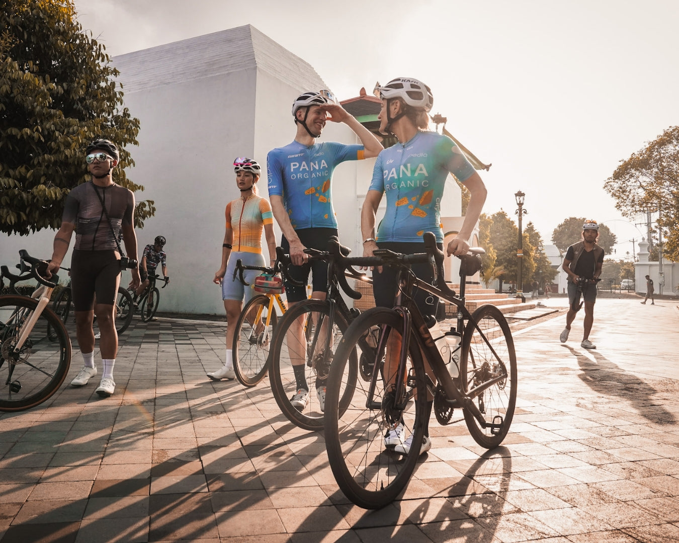 Two cyclists in Pana Organic Pedla kit standing next to their bikes as the sun shines behind them