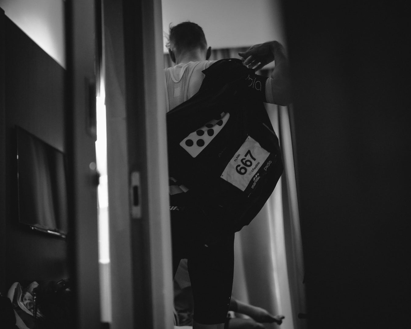 A cyclist gets ready for the cycling race by putting their race jersey on in the hotel room, black and white image