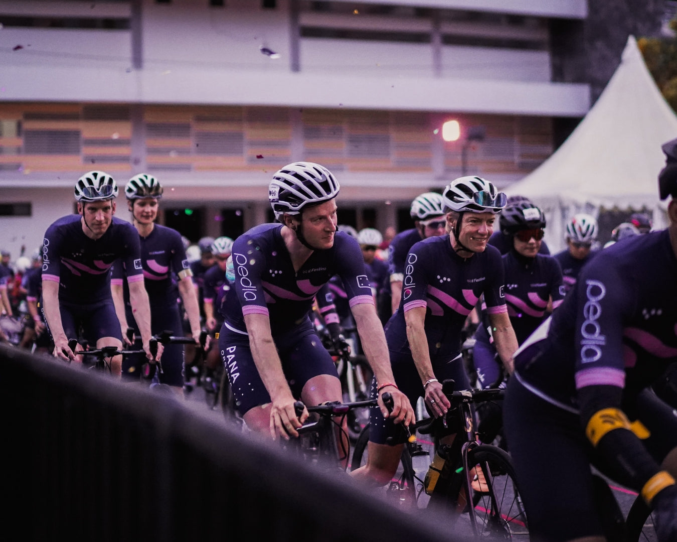 Cyclists amongst a larger group of cyclists waiting in the marshalling area at dawn before the race
