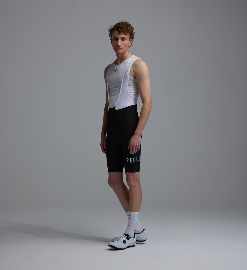 PEDLA Heritage men's training cycling bib shorts (side view) in black with white breathable mesh bib, compressive fit, premium European fabrics, updated brace system, seamless cuff, reflective logos and supportive chamois