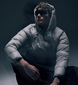 Close-up of model wearing PEDLA x N-0 grey lightweight insulated down pullover with DWR nylon shell, channel-quilted baffles, insulated hood with collar zip, zipped hand pockets, elastic cuffs and drawcord hem, technical unisex cycling streetwear