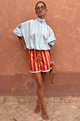 Blue Stripe Batwing Blouse with a high tie neck and shirred cuffs, styled with red shorts against a terracotta wall.