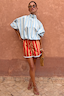 Blue Stripe Batwing Blouse with a high tie neck and shirred cuffs, styled with red shorts against a terracotta wall.