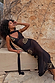 Petite Black & Gold Simona Maxi Dress in sheer mesh with gold sequins and a multi-wear scarf, styled against a stone wall.