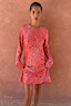 Red Spliced Fawn Mini Nola Tie Dress featuring a white speckled print and long sleeves against a terracotta wall.