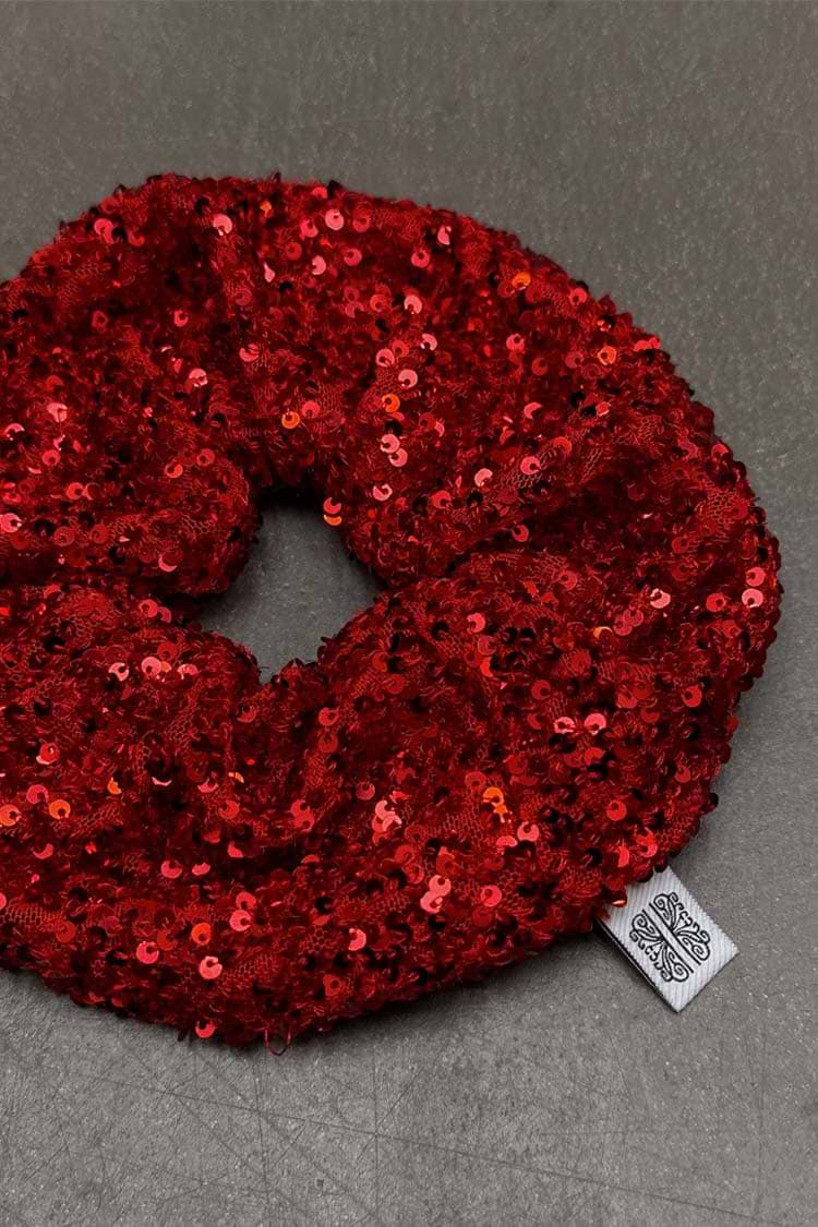Red Sequin Scrunchie with shimmering red sequins and a signature white brand logo tag on a grey surface.